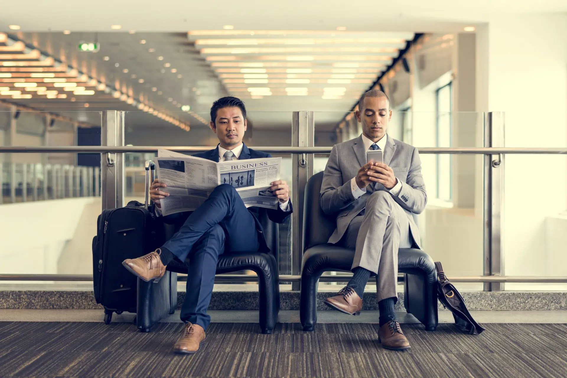 Two men who have crossed their legs reading the newspaper and checking their mobile phone